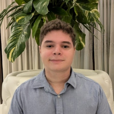 Leandro Gabriel sorrindo, de camisa azul, sentado numa poltrona branca com uma planta ao fundo